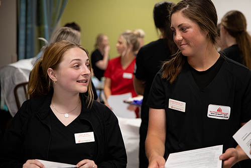 two young women in nursing student scrubs listen and respond to an instructor off camera