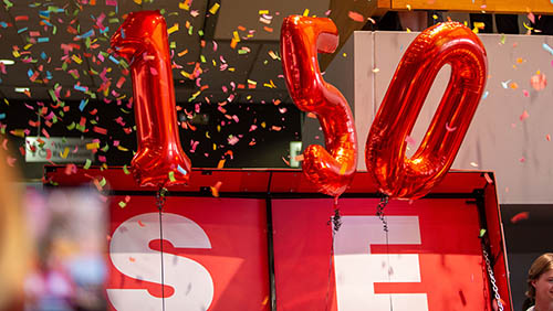 a large birthday present is open and 150 balloons have risen out of it. confetti is raining down