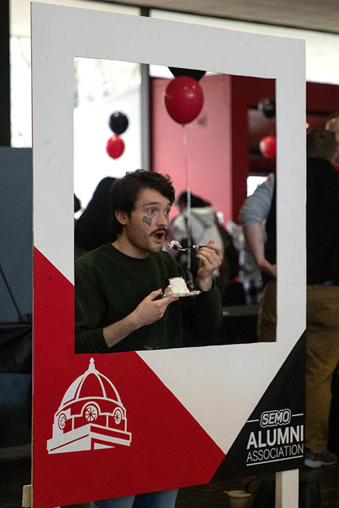 a student holds a plate of cake and is in the process of taking a bite, and posing for a photo with a prop frame