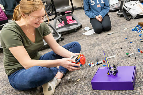 a female education student is seated on the floor with lego bricks and wiring scattered around her. she's holding a construction made of lego and laughing