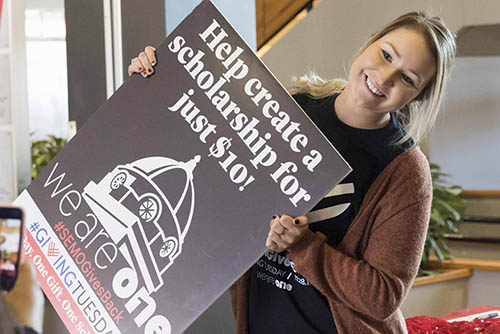 a young woman holding a sign advertising supporting a scholarship for ten dollars poses for a phone photograph