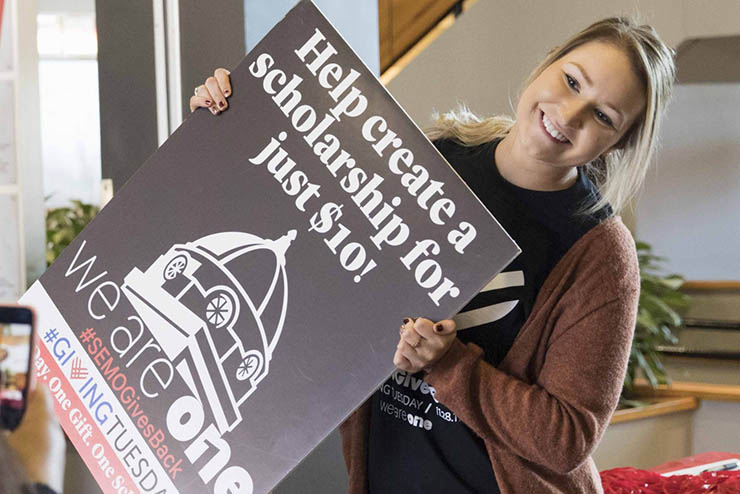 a young woman holding a sign advertising supporting a scholarship for ten dollars poses for a phone photograph