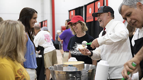 the university president and his wife wearing aprons and hats serve ice cream to students while talking and laughing