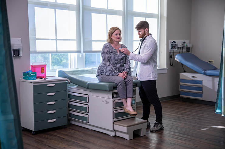 a nursing student in a white coat is standing in an exam room with an older person on the exam bench listening to her chest with his stethoscope