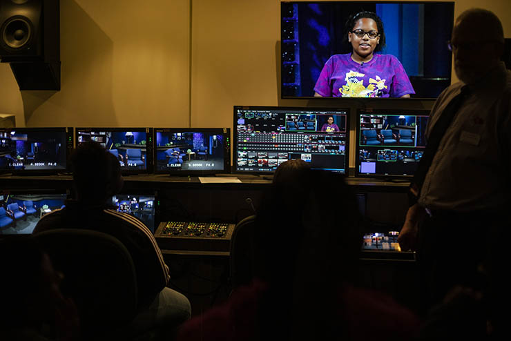 students sit in the dark in a production room managing control boards and watching the production on a screen on the wall