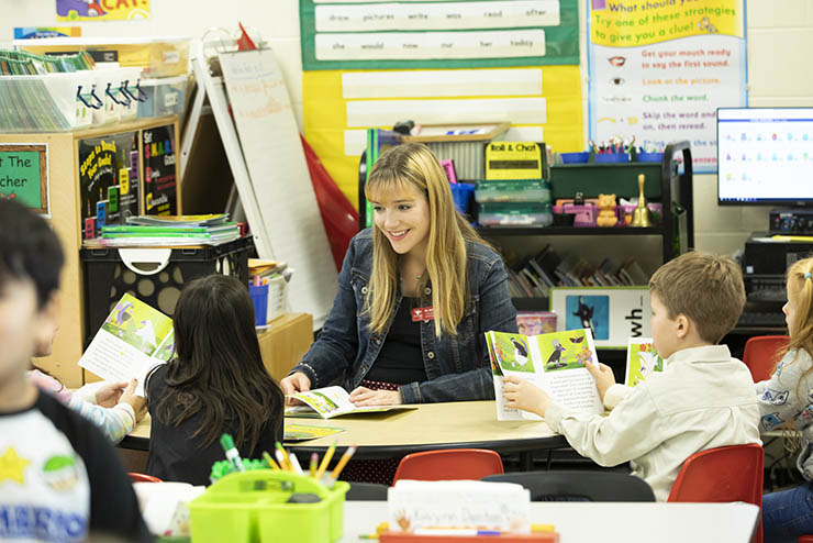 a young student teacher sits in front of a classroom of small children reading to them