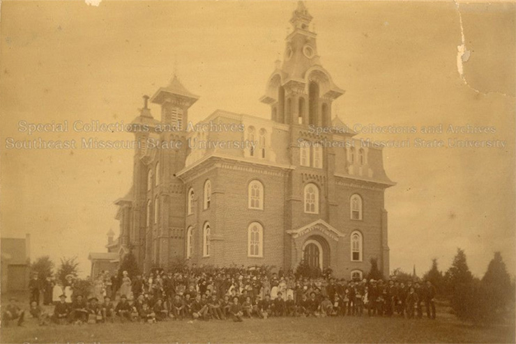 Photo from 1870s of Normal School, SEMO's original school building. It has gothic architecture and has people sitting in front of it.