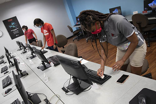 students in a computer lab wearing face masks over their noses and mouths wipe down keyboards with sanitizing wipes