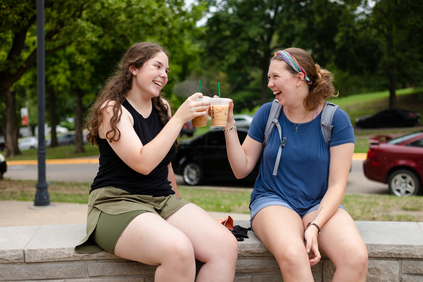 Two students sitting outside on campus doing a "cheers" pose with their starbucks drinks