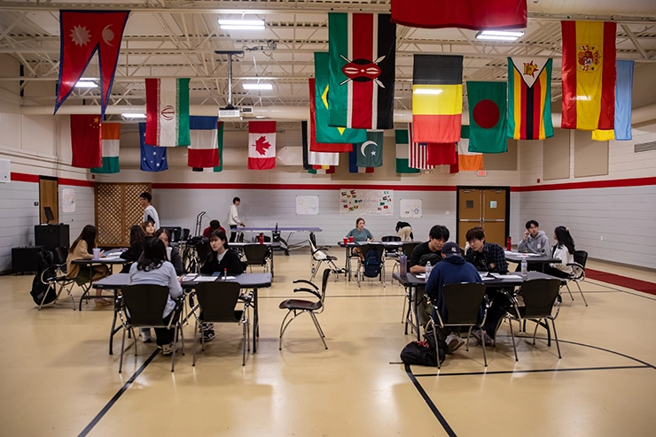 International students meet at the international student meet and greet at desks in a room with international flags hanging from the ceiling. 