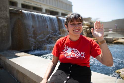 A student in a SEMO t-shirt smiles and waves while sitting on the edge of a fountain.