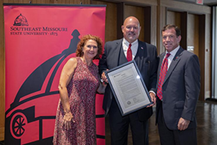 Members from the Board of Regents stand by a red banner with a black dome supergraphic and university logo.  