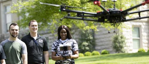 Students wait their turn as one student flies a drone on campus outside on a sunny spring day.