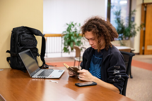 A student reads a book while working on his laptop.