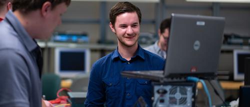 A student smiles while working on a computer surrounded by wires and computer parts.