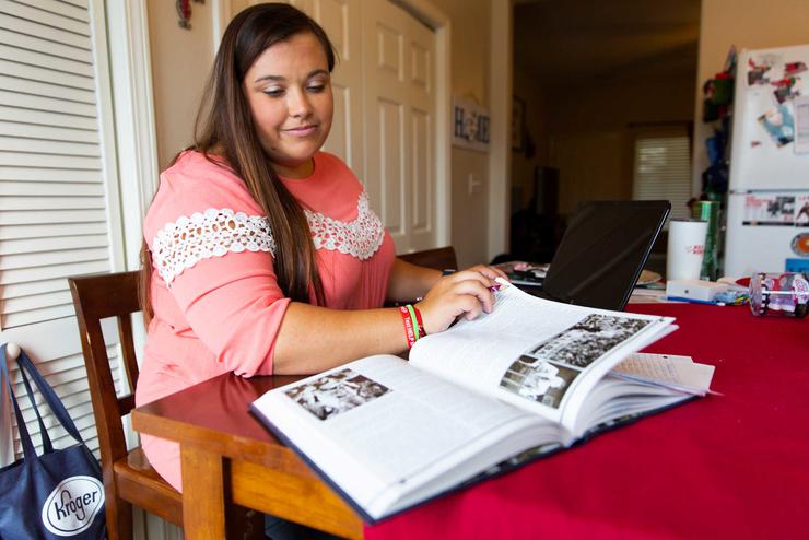 A student sits at a table and reads a book while working on a laptop.