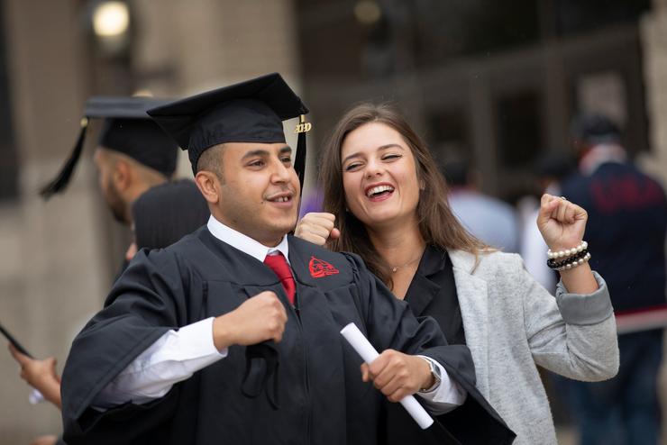 A graduate, dressed in cap and gown, holds a diploma and cheers with a peer.