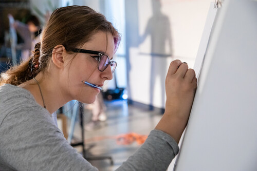 A student holds a pencil in her mouth while drawing on an easel.