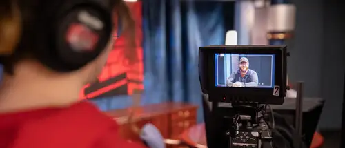 a media management degree major stands behind a television camera wearing a headset and watching what is going on on the set as a part of the media management course