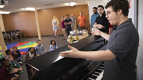 a instrumental music education degree student stands at a piano and addresses a class of small children