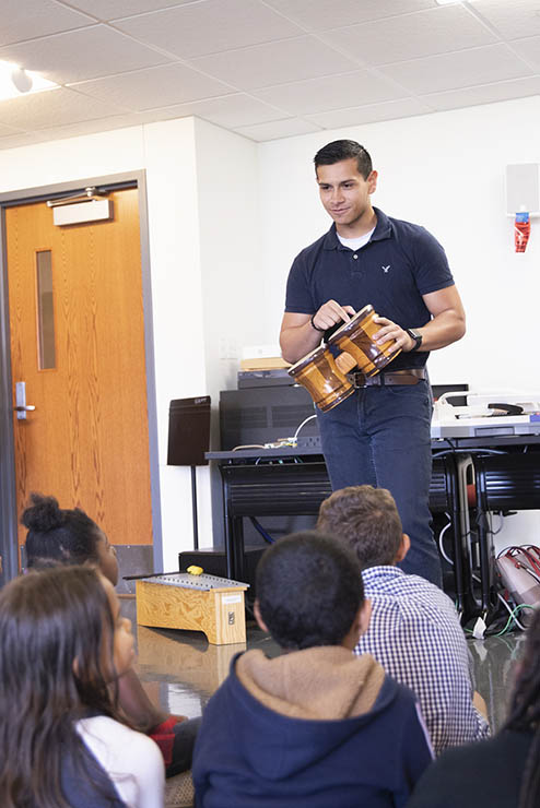 a student in the instrumental music programs stands in front of a class of small children and explains how to play a hand drum
