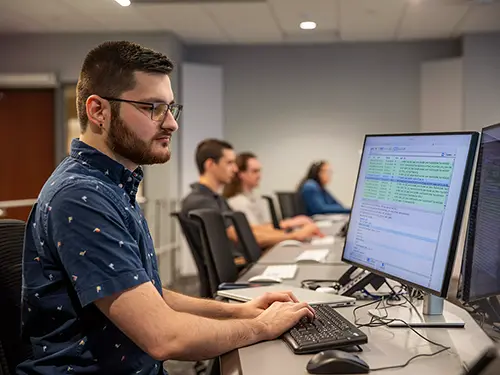 a cybersecurity major student sits at a table in the cyber range working on his laptop intently