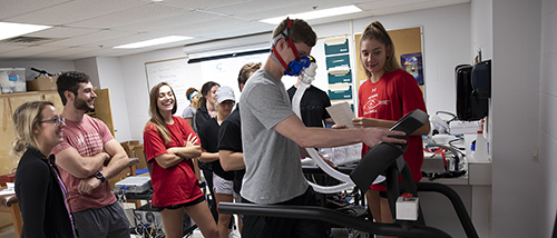 Three students pursuing a health and exercise science degree and their teacher stand around a treatment table working with equipment in the physical therapy room. 