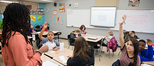 middle school education social science stands at the front of a classroom of young students teaching as they raise their hand to answer her question. 