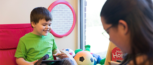 A young exceptional needs child wearing a bright green shirt smiles as he looks at a book a cross categorical teacher in the cross categorical special education program is showing him