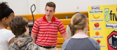 A family and consumer science major talks to several students in a gymnasium at a local school about nutrition information. 