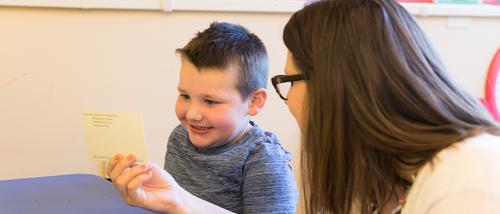 A child at the Autism Center smiles happily as a childcare and guidance associates degree student hold up a card for him to look at.