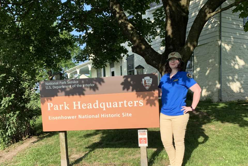Kelsey Barnett, historic preservation degree alumni, wearing a blue shirt, khaki pants, and a hat, smiles beside the sign for the Eisenhower National Historic Site.