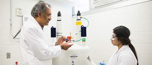 A student wearing protective eyewear and a lab coat takes notes as her professor demonstrates an experiment.