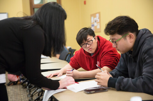Two students listen to a teacher who is leaning over them explaining work.