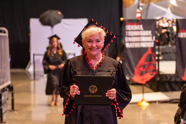 an older, non traditional student happily shows off her diploma at graduation ceremonies