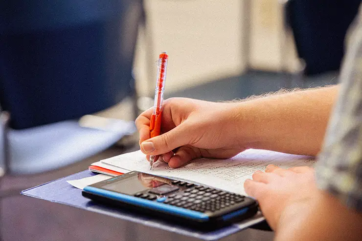 a student works on math equations in a notebook with a calculator beside them
