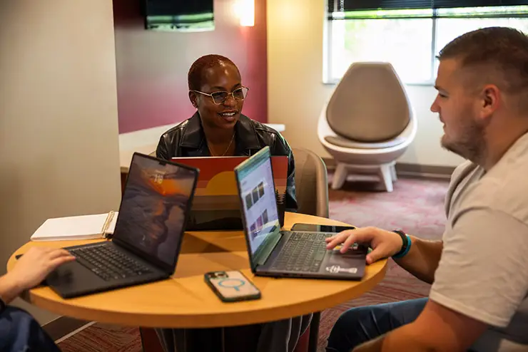 three students sit together at a table with their laptops in front of them
