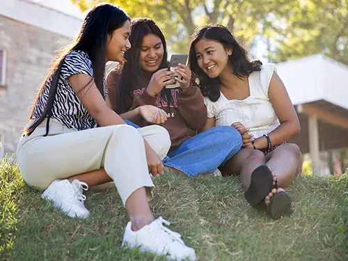 a group of three SEMO international students sit together in the grass and look at a phone screen
