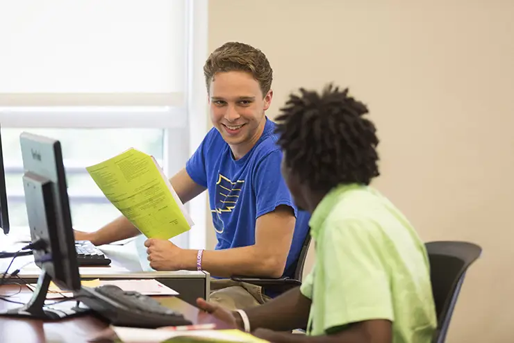 two students sit together in a computer lab and discuss their work