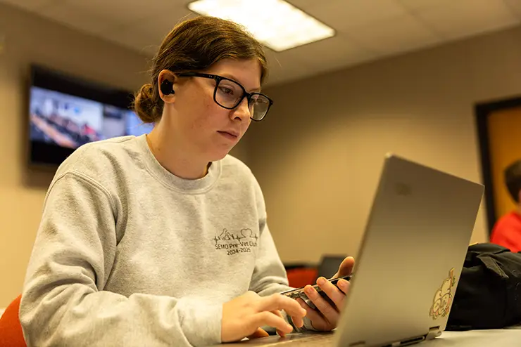 a SEMO student sits at her laptop, headphones in her ears and her phone in her hand, doing research