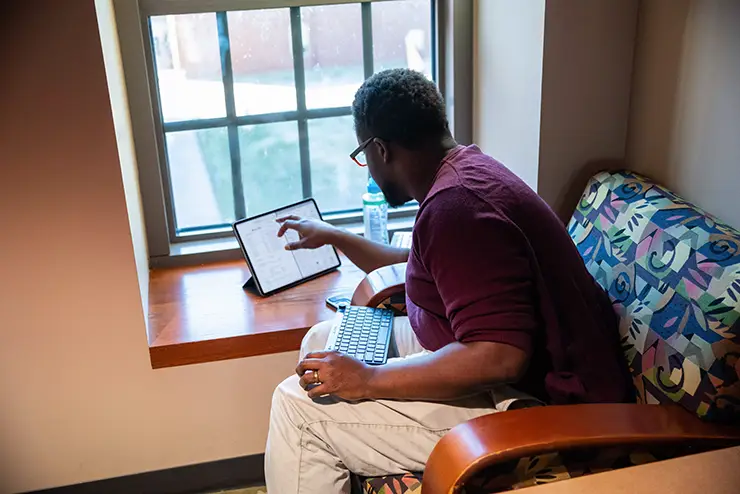 a student sits at a window with his tablet, working on the screen