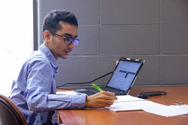 a student sits at a study table with his laptop open taking notes in a notebook
