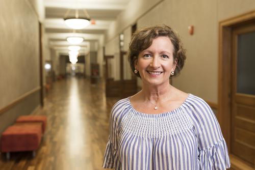 Wearing a blue and white striped blouse, Marie Gill smiles as she stands in Academic Hall. 