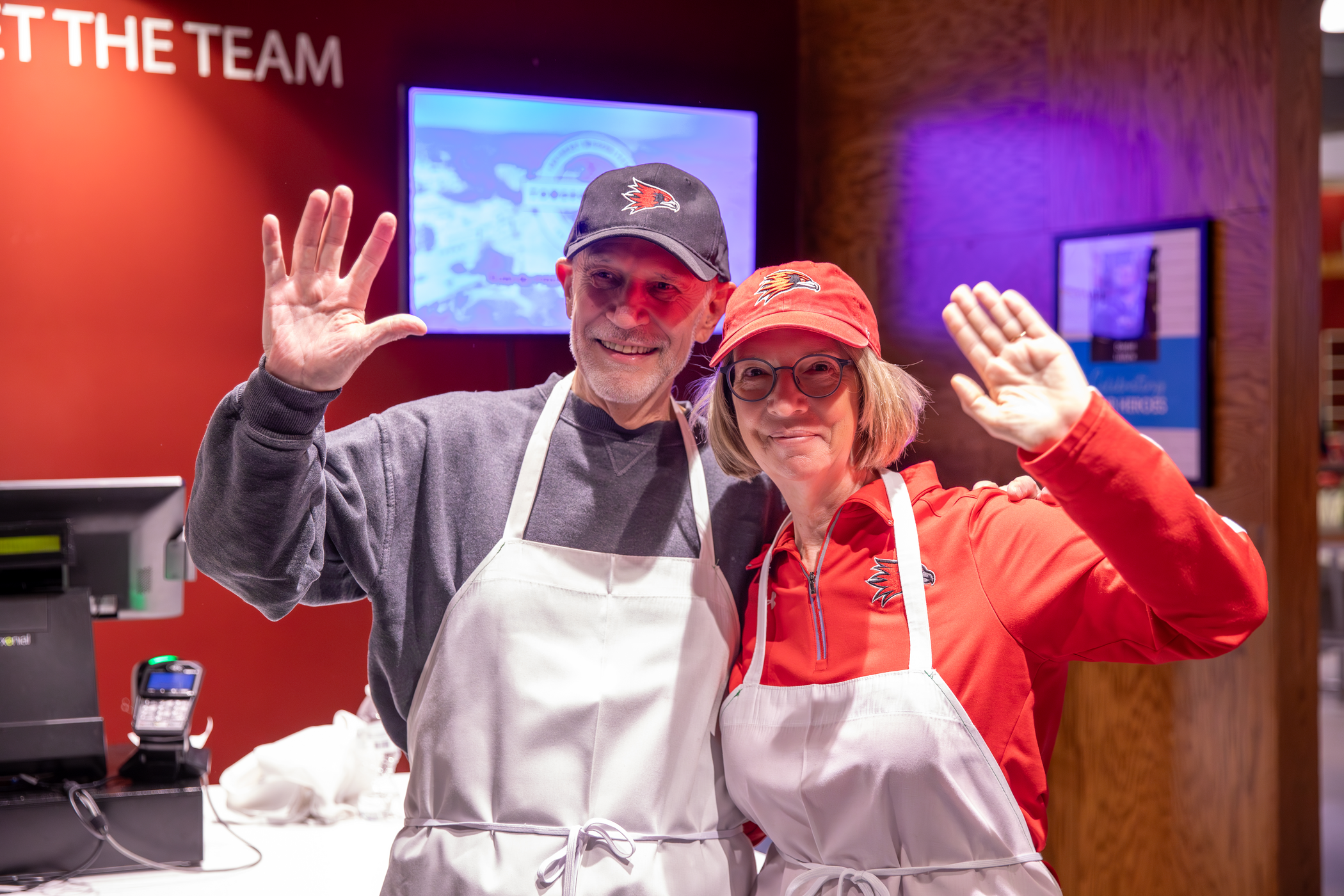 Dr. Vargas and Pam smiling and waving at the camera before serving students at Late Night Breakfast, creating a warm and welcoming atmosphere.