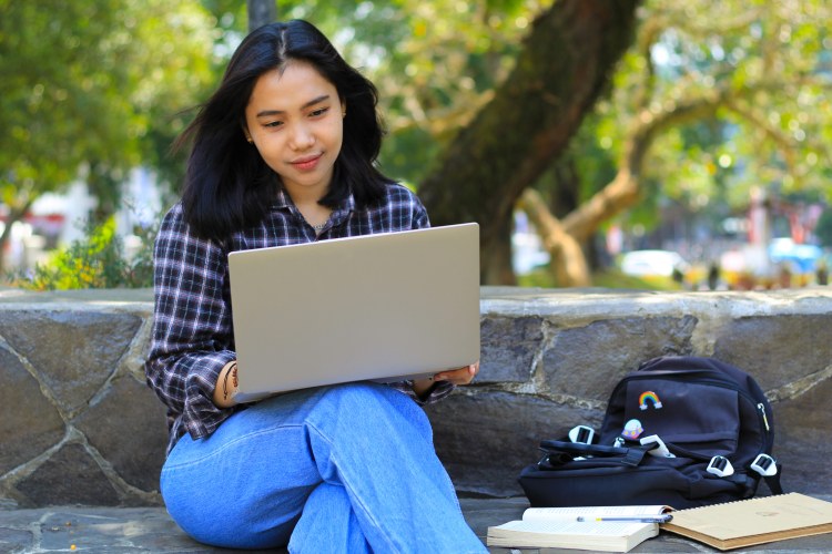 An online college student pursuing stackable credentials sitting outside holding a laptop.