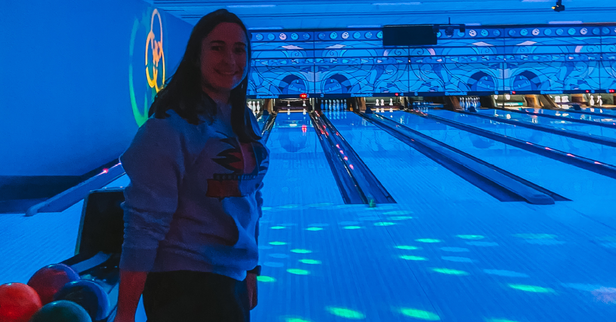 A woman smiles while holding a bowling ball in front of lanes. The bowling alley is lit with purple light for a glow bowl.