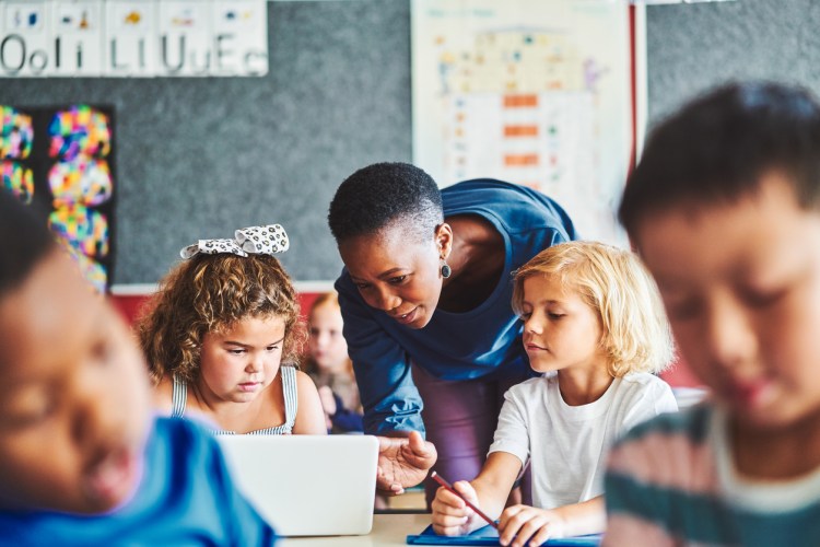 A teacher helping a young student use a technology device in a classroom.