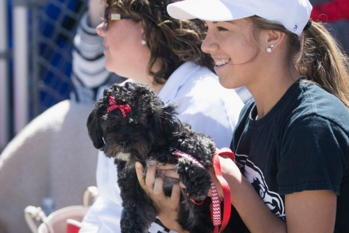 A female student wearing a white hat smiles while holding a black dog with a red bow in its hair.