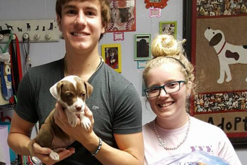 A male and female student smile while holding a small puppy.