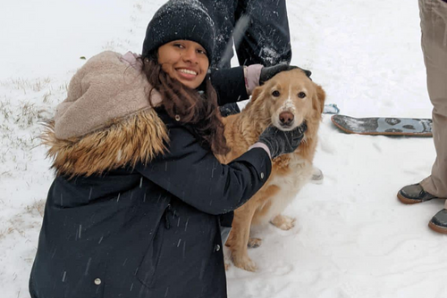 A student wearing a large jacket smiles next to a dog in the snow.